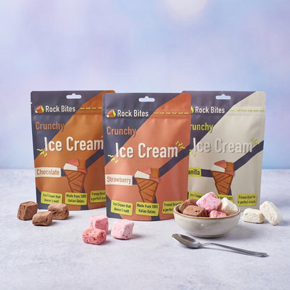 Three packages of Rock Bites Crunchy Ice Cream with a bowl of ice cream and freeze dried ice cream on a light background
